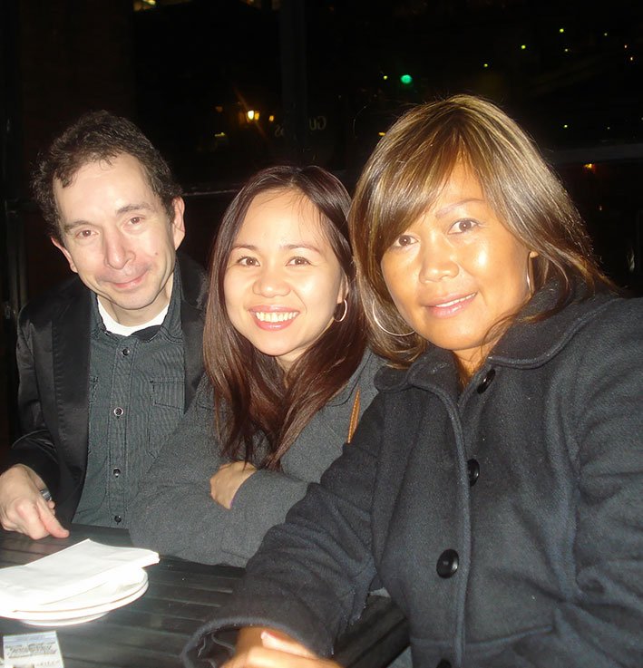 Three guests sit side by side at a table during the fundraising event, dressed in coats and smiling toward the camera.