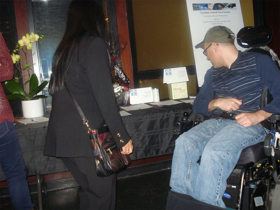 Adult using a power wheelchair looks over a silent auction table covered with bid sheets and gift baskets, while another guest stands nearby reading the display.