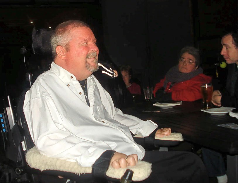 A man using a power wheelchair sits at a table at the fundraising event, facing other guests across the table.