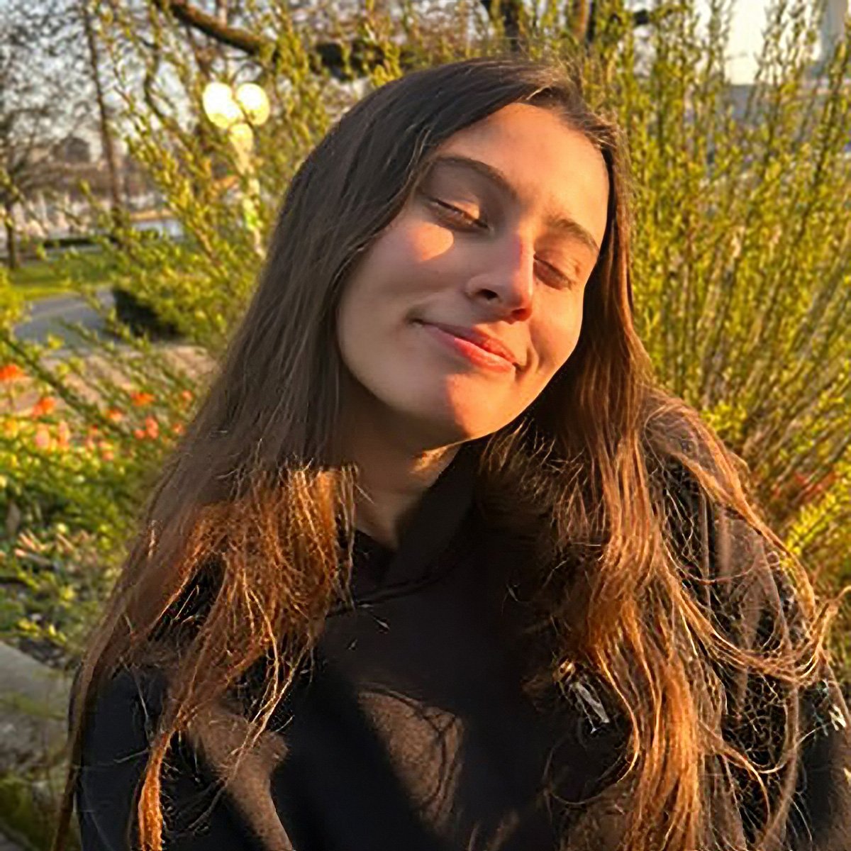 Jessica Demers sits outdoors in soft golden-hour light, wearing a dark hoodie with green shrubs and a park setting blurred in the background.